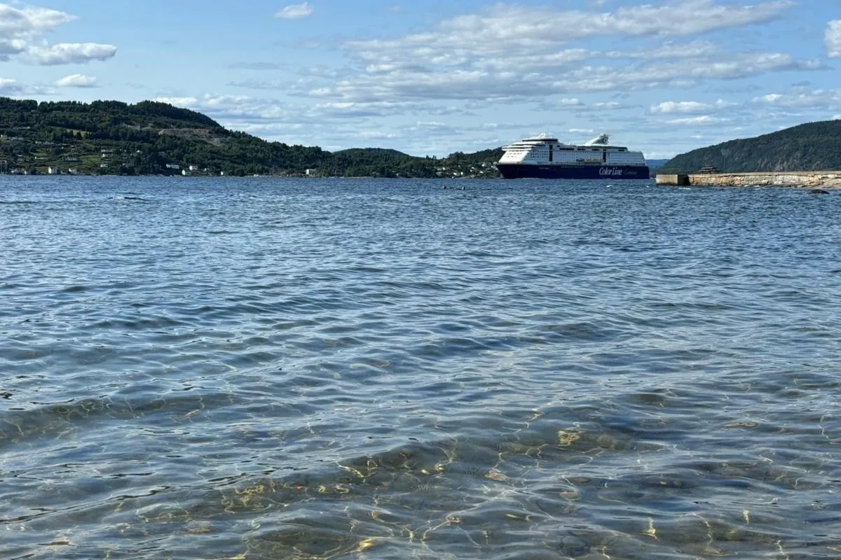 Un traghetto di Color Line sul fiordo di Oslo, visto da una riva con l’acqua limpida e bassa, tra colline coperte di boschi
