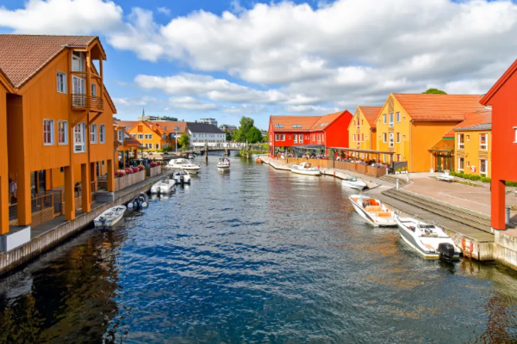 Case di legno arancioni e rosse lungo un canale a Kristiansand, con piccole barche a motore ormeggiate sotto un cielo azzurro con nuvole