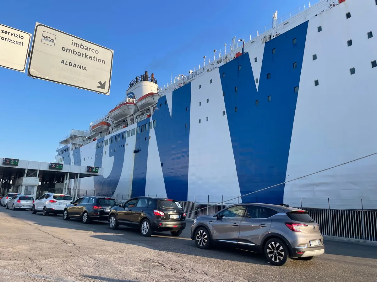 Auto in fila per l’imbarco nel porto di Bari, sotto un cartello con imbarco embarkation Albania, accanto a una grande nave bianca e blu di GNV