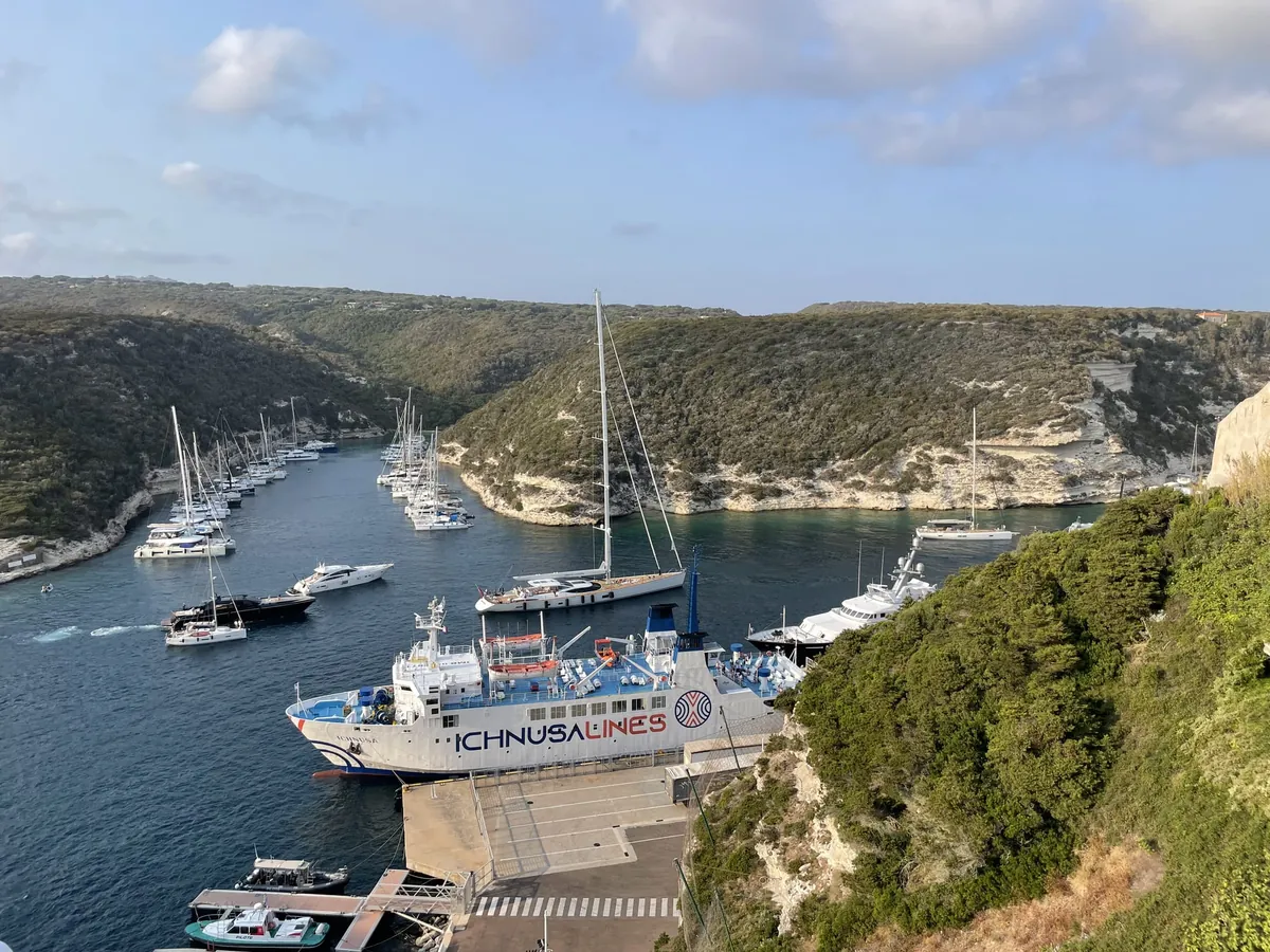Il traghetto bianco Ichnusa ormeggiato nel porto di Bonifacio, visto dall’alto, in una stretta insenatura tra scogliere calcaree bianche coperte di verde, con decine di barche a vela e a motore sull’acqua