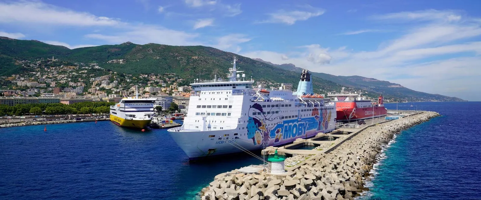 Il porto di Bastia visto dal mare, con tre traghetti in banchina: uno giallo di Corsica Ferries, il bianco Moby Orli con i personaggi dei Looney Tunes e uno rosso di Corsica Linea, davanti alla città e alle montagne
