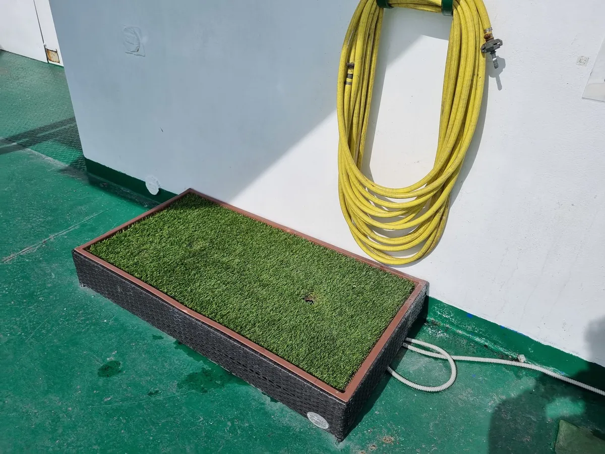 Una vaschetta rettangolare di erba sintetica con cornice nera su un ponte esterno verde, con una canna dell’acqua gialla arrotolata sulla parete bianca sopra