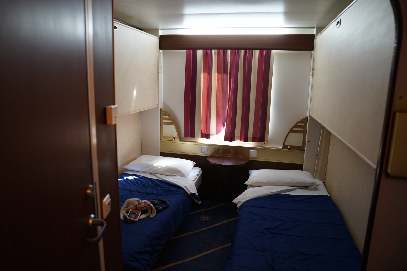 Cabina da quattro con due letti bassi dai copriletto blu, due cuccette ribaltate contro le pareti e un oblò con una tenda a righe rosse