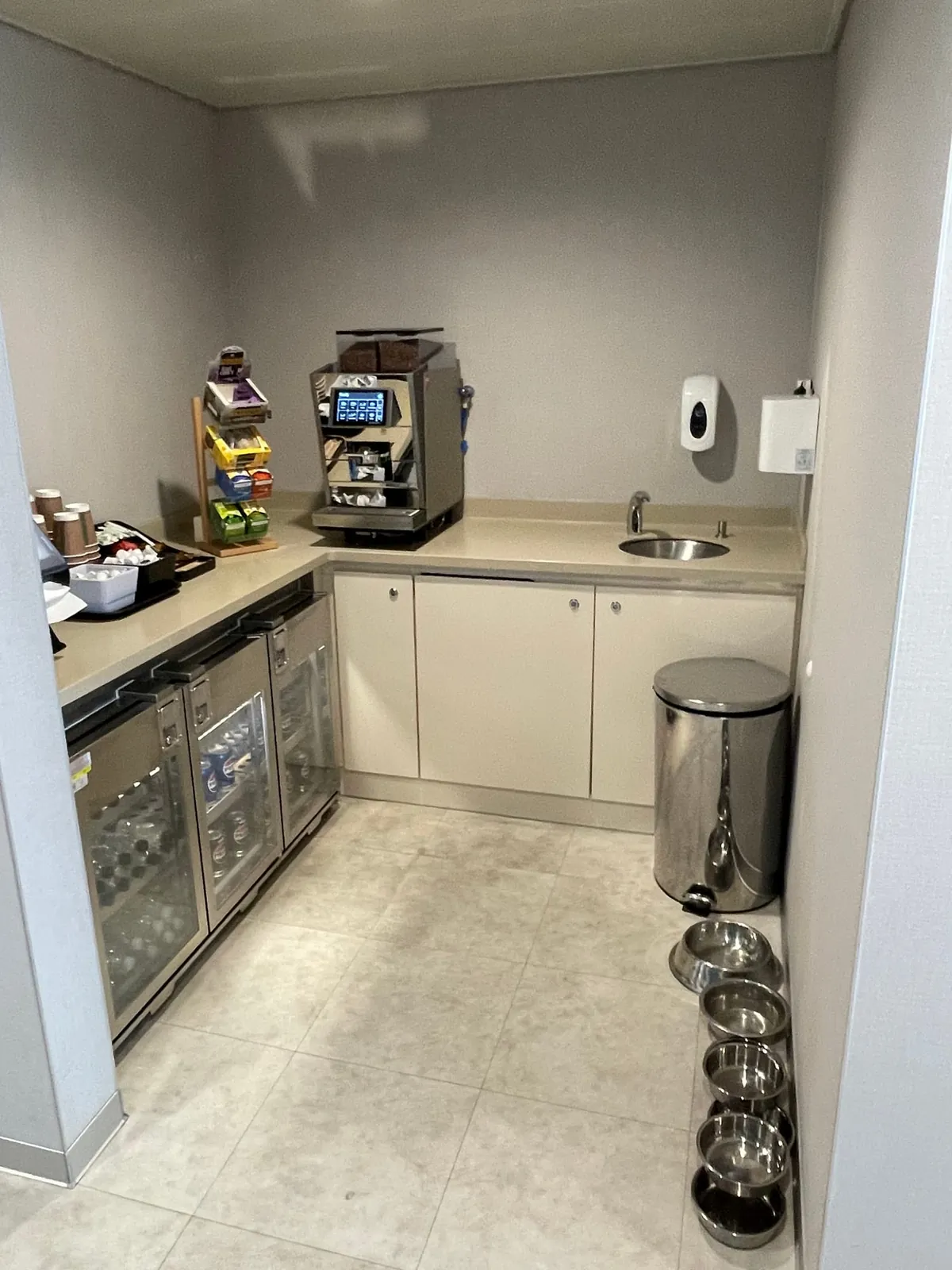 Un angolo cucina con una macchina del caffè, frigoriferi con lattine e bottiglie, bicchieri e snack sul bancone, un lavello, un cestino e sul pavimento una fila di ciotole in acciaio