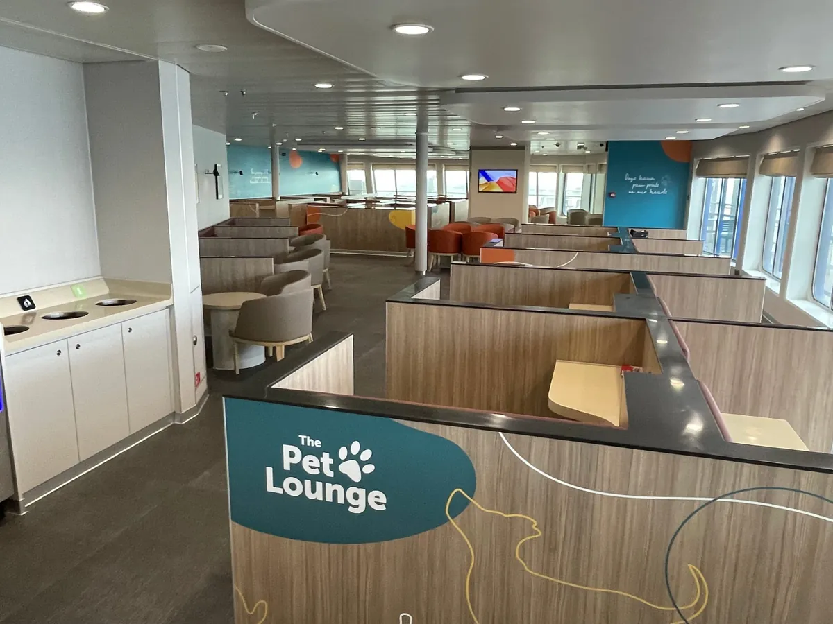 The Pet Lounge a bordo: box separati da pareti di legno, sedie e tavolini rotondi, pareti blu petrolio con la scritta Dogs leave paw prints on our hearts e finestre su un lato