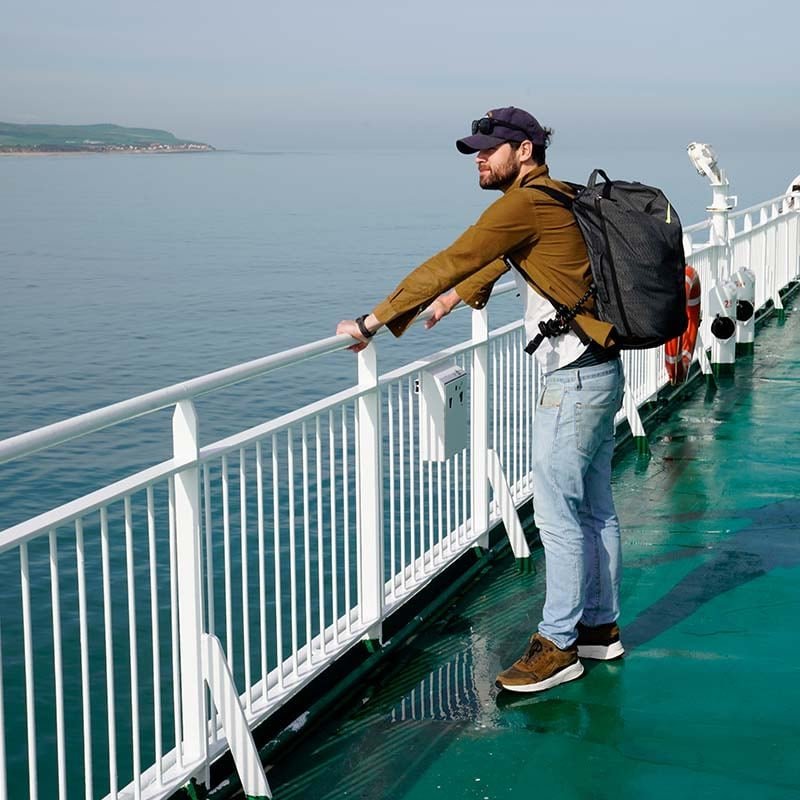 Rob Tigchelaar, con un cappellino e gli occhiali da sole sulla visiera e uno zaino in spalla, appoggiato al parapetto bianco di un ponte verde, guarda un mare calmo con una costa in lontananza